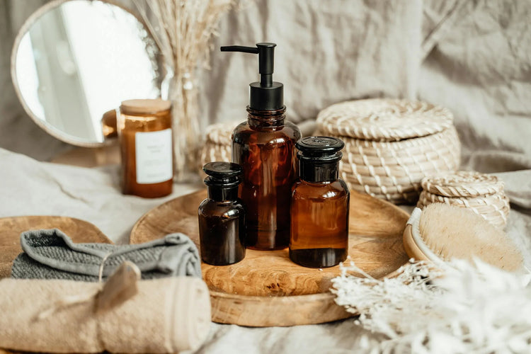 A bathroom setup with amber glass jars, cotton towels, wooden trays, woven baskets, a natural candle, and bamboo brush, highlighting items made from natural materials.